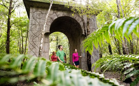 People walking through a large monument sitting between trees and bush on the Kaitoke regional park trail.