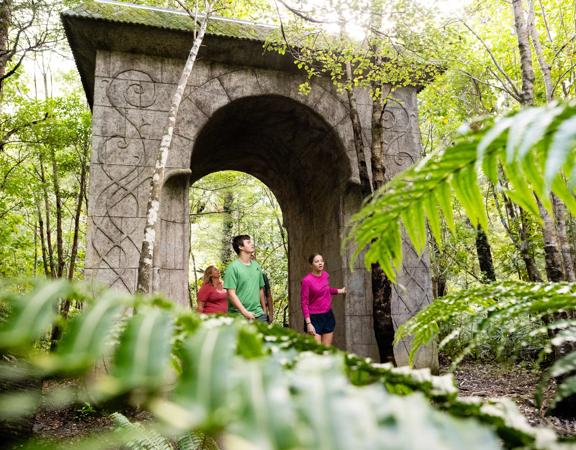 People walking through a large monument sitting between trees and bush on the Kaitoke regional park trail.