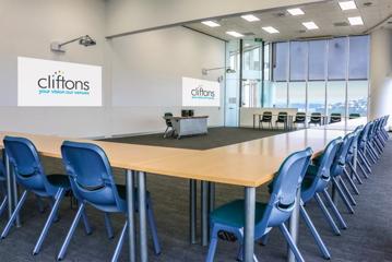 Interior of a cliftons conference room, with 20 chairs around 8 tables in a square facing a projector.