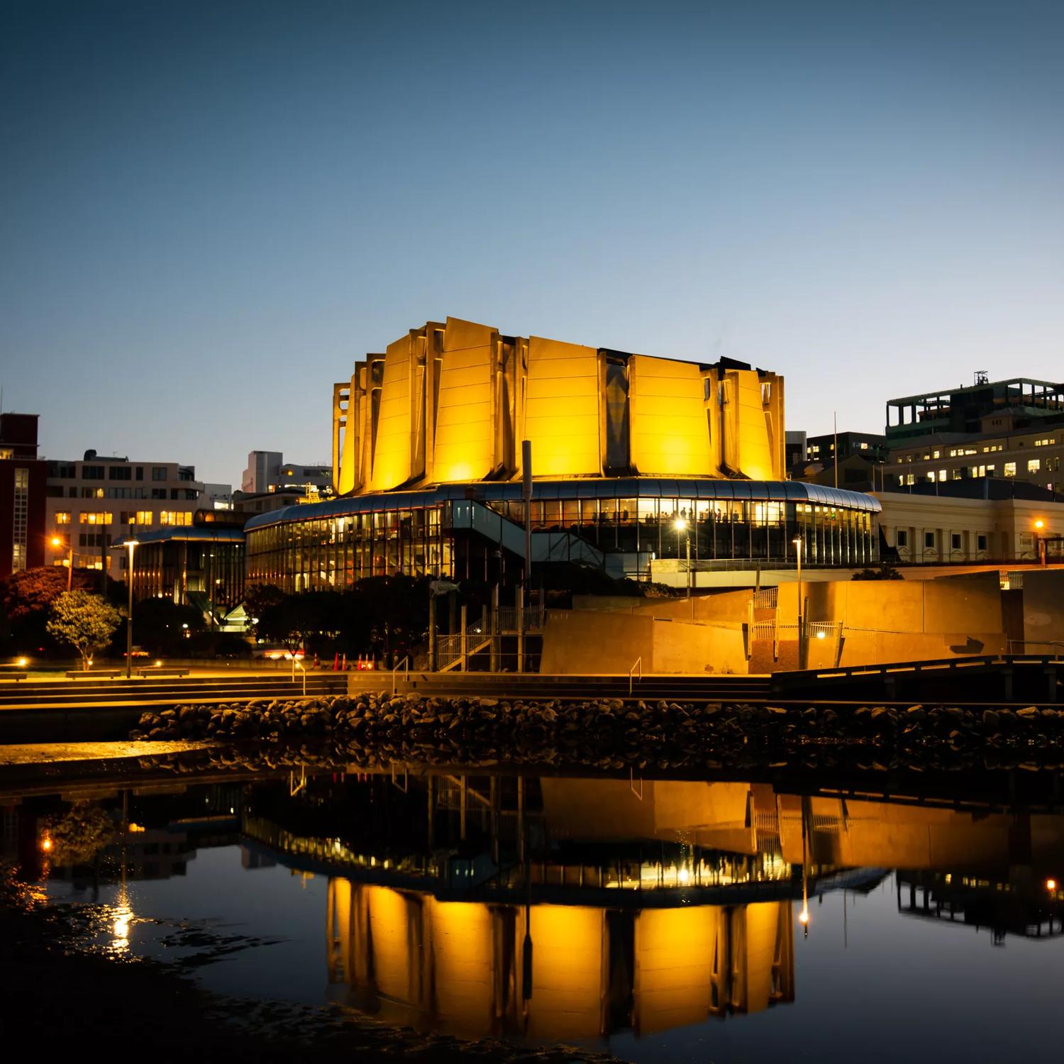  Michael Fowler Centre lit up in celebration of the Prime Minister of New Zealand Jacinda Ardern's announcement of a baby girl. with bright yellow lights reflected onto the waterfront and into the sky at nighttime.