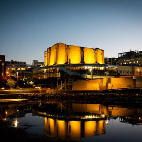 Michael Fowler Centre lit up in celebration of the Prime Minister of New Zealand Jacinda Ardern's announcement of a baby girl. with bright yellow lights reflected onto the waterfront and into the sky at nighttime.