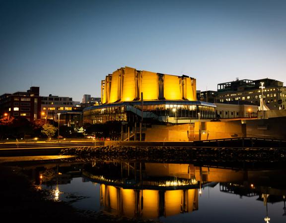 Michael Fowler Centre lit up in celebration of the Prime Minister of New Zealand Jacinda Ardern's announcement of a baby girl. with bright yellow lights reflected onto the waterfront and into the sky at nighttime.