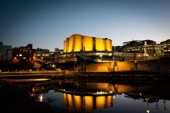 Michael Fowler Centre lit up in celebration of the Prime Minister of New Zealand Jacinda Ardern's announcement of a baby girl. with bright yellow lights reflected onto the waterfront and into the sky at nighttime.