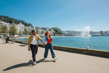 Two people stroll along Oriental Bay Parade in Wellington holding hands on a sunny day.