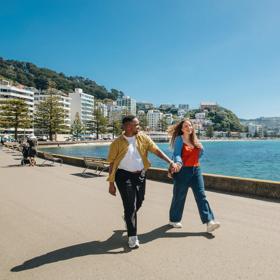 Two people stroll along Oriental Bay Parade in Wellington holding hands on a sunny day.