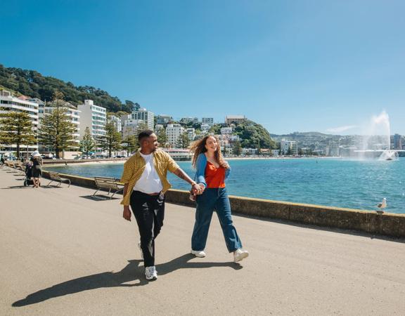 Two people stroll along Oriental Bay Parade in Wellington holding hands on a sunny day.