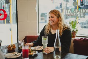 A smiling person sits at a booth at Chaat Street, a tapas-style Indian restaurant in Wellington, enjoying food and drinks.