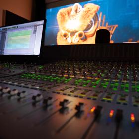 A close-up of a sound control desk with lots of buttons and lights. Two screens are in the background.