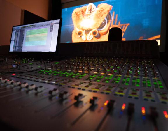 A close-up of a sound control desk with lots of buttons and lights. Two screens are in the background.