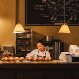 A pastry chef in a white coat and grey apron looks down while making cheese scones at Pravda Café & Grill. There are two racks of scones cooling on racks on the counter.