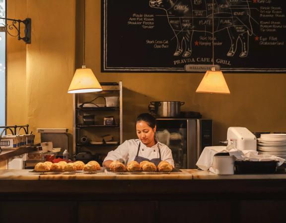 A pastry chef in a white coat and grey apron looks down while making cheese scones at Pravda Café & Grill. There are two racks of scones cooling on racks on the counter.