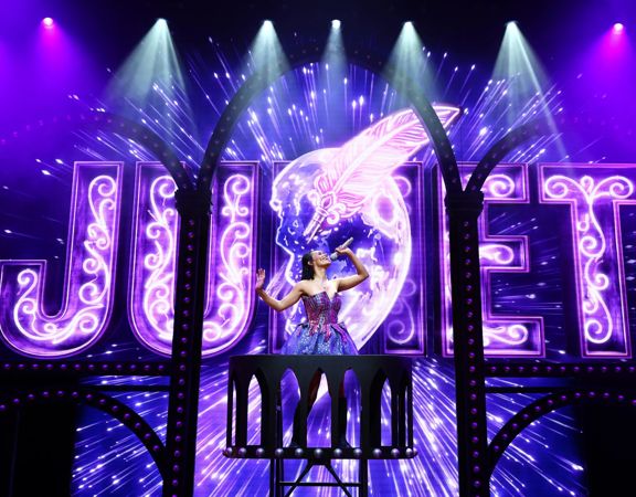 The lead actress in & Juliet sings into a microphone on an elevated platform.