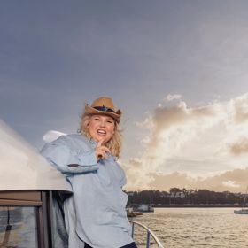 Comedian, Urzila Carslon, stands on the edge of a boat wearing a cowboy hat.