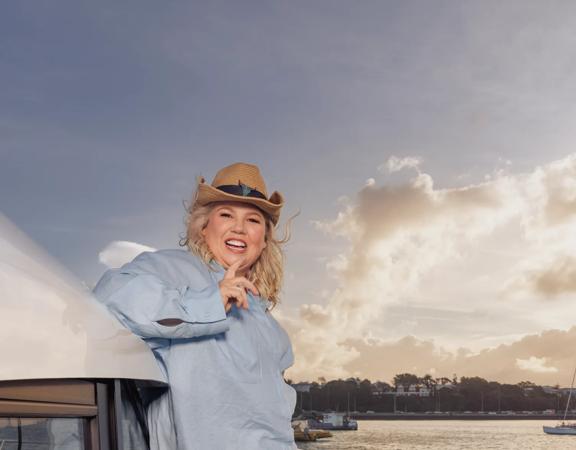 Comedian, Urzila Carslon, stands on the edge of a boat wearing a cowboy hat.