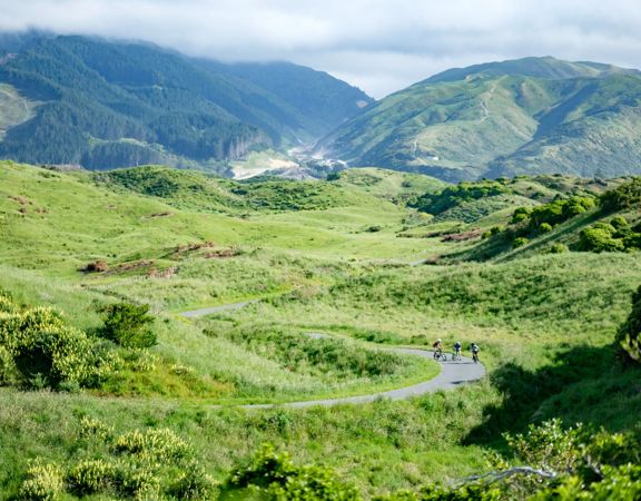 A wide shot of three cyclists on a winding path in a grassy valley in Queen Elizabeth Park.