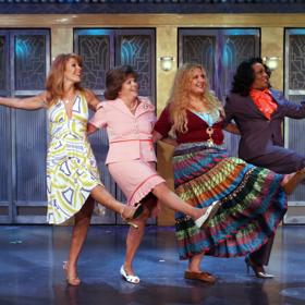 Four joyful actresses line up on stage, kicking their right legs high in a spirited can-can routine.