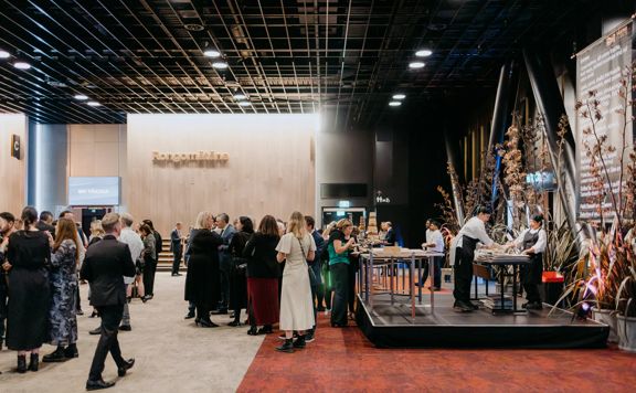 Inside Tākina Wellington Convention & Exhibition Centre at the opening event. There is food catering and many people are mingling in formal wear.