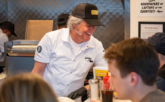 Chef in a white uniform and cap serving burgers behind a busy diner-style counter, engaging with customers as plates, condiments, and drinks sit in the foreground.