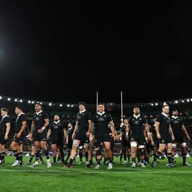 New Zealand's national rugby union team the All Blacks wearing black rugby uniforms, standing in the middle of a stadium at night.