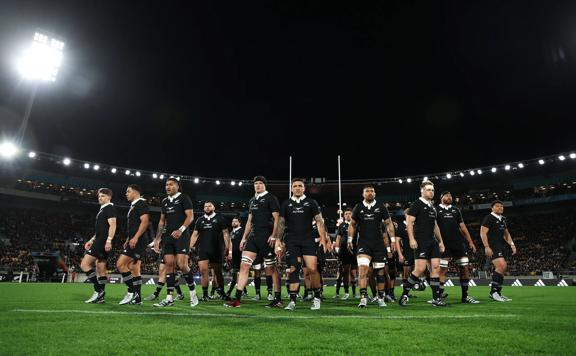 New Zealand's national rugby union team the All Blacks wearing black rugby uniforms, standing in the middle of a stadium at night.