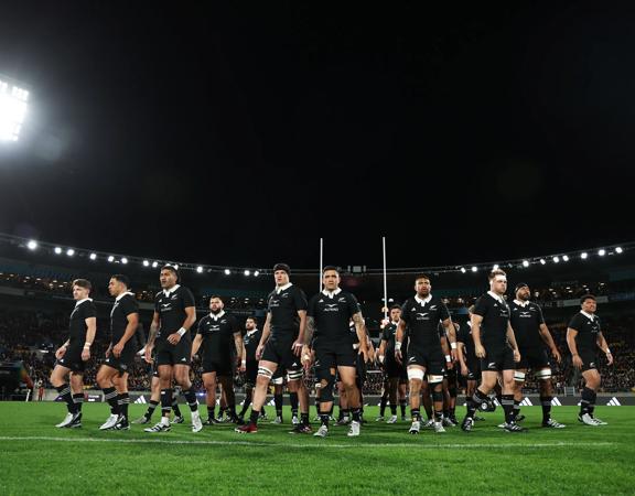 New Zealand's national rugby union team the All Blacks wearing black rugby uniforms, standing in the middle of a stadium at night.