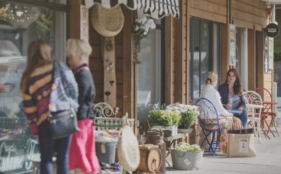 People sitting outside the Kāpiti Raumati shops, with flowers, veggies and vintage chairs outside.