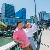 Two people are standing on Kumutoto Wharf near Lambton Quay on a sunny day. The water behind them is green and the sky is blue.