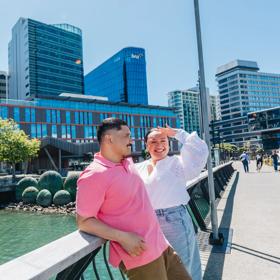 Two people are standing on Kumutoto Wharf near Lambton Quay on a sunny day. The water behind them is green and the sky is blue.