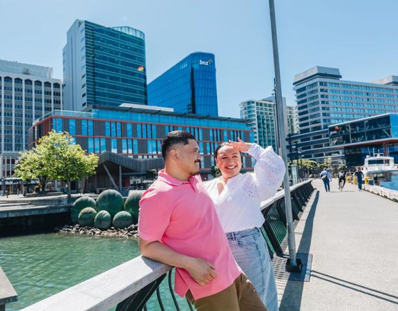 Two people are standing on Kumutoto Wharf near Lambton Quay on a sunny day. The water behind them is green and the sky is blue.