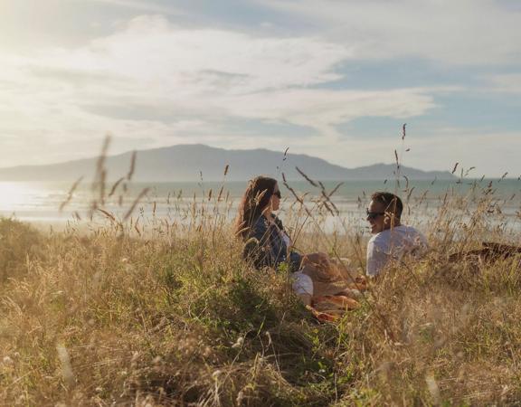 Two people sit in tall grass and admire a scenic vista with Kapiti Island in the background.