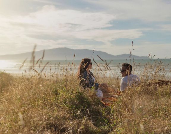 Two people sit in tall grass and admire a scenic vista with Kapiti Island in the background.