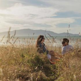 Two people sit in tall grass and admire a scenic vista with Kapiti Island in the background.
