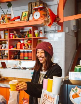 A smiling barista wearing a red knitted hat hands a takeaway coffee to a customer.