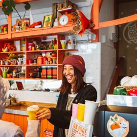 A smiling barista wearing a red knitted hat hands a takeaway coffee to a customer.