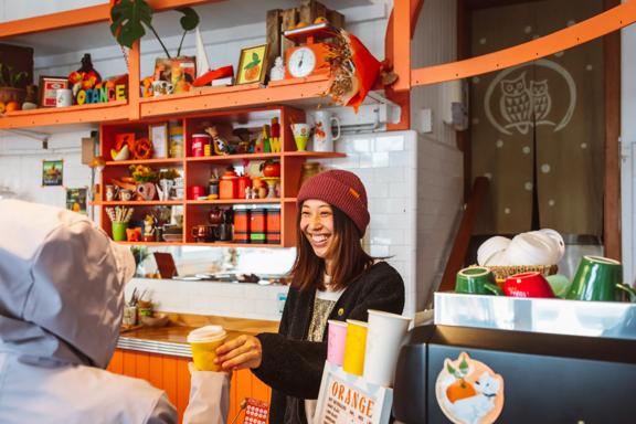 A smiling barista wearing a red knitted hat hands a takeaway coffee to a customer.