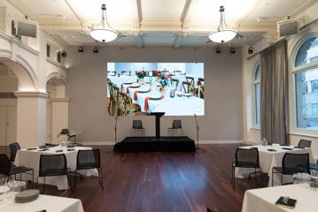 conference room with hard wood floors, 4 tables each with 4 chairs, facing towards a projector in the centre of the room and podium.