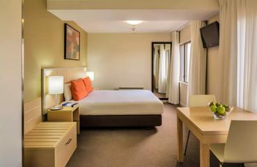 A guest room at the Travelodge Hotel has a bed against the wall with crisp white sheets, and a small table with a bowl of fruit.