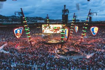 A stadium full of people shown at dusk, the stage is in the centere of the stadium, surrounded by bright LED lights and screens.