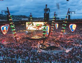 A stadium full of people shown at dusk, the stage is in the centere of the stadium, surrounded by bright LED lights and screens.