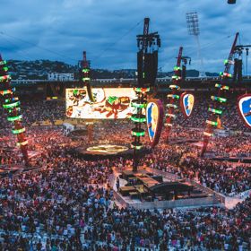 A stadium full of people shown at dusk, the stage is in the centere of the stadium, surrounded by bright LED lights and screens.