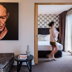 Inside a room at the Museum Apartment Hotel in Te Aro, Wellington. The room features a modern aesthetic, with a neutral colour palette of grey, beige and gold accents. A person is walking towards the window. 