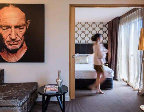 Inside a room at the Museum Apartment Hotel in Te Aro, Wellington. The room features a modern aesthetic, with a neutral colour palette of grey, beige and gold accents. A person is walking towards the window. 