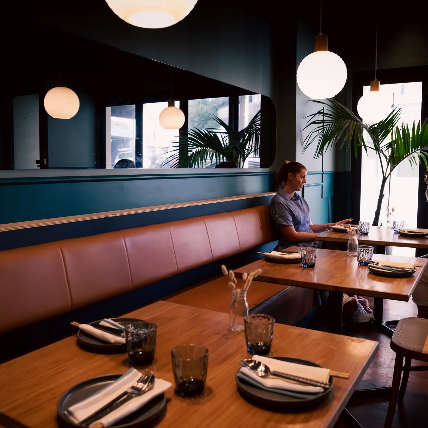 Two customers are are eating at a table in Koji. Lantern lights hang above them, and a fern sits to their left.