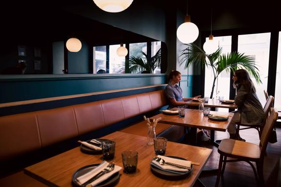 Two customers are are eating at a table in Koji. Lantern lights hang above them, and a fern sits to their left.