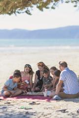 A family of eight sit and eat food on a picnic blanket under a shady spot on a sandy beach.