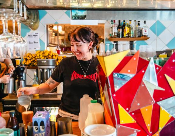 A waiter inside Seashore Cabaret is pictured smiling while pouring a coffee. Vibrant colours decorate the room.