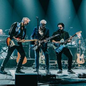 Three guitarists from the British rock band 10cc perform on stage with supporting keyboards and drums.