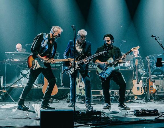 Three guitarists from the British rock band 10cc perform on stage with supporting keyboards and drums.