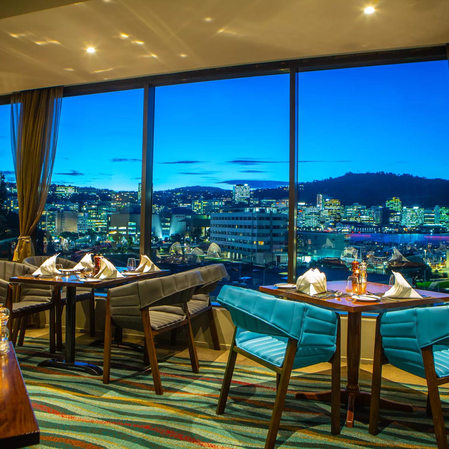 The Copthorne hotel dining room, chairs and tables overlook Wellington city at sunset through floor to ceiling windows.