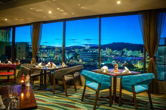 The Copthorne hotel dining room, chairs and tables overlook Wellington city at sunset through floor to ceiling windows.